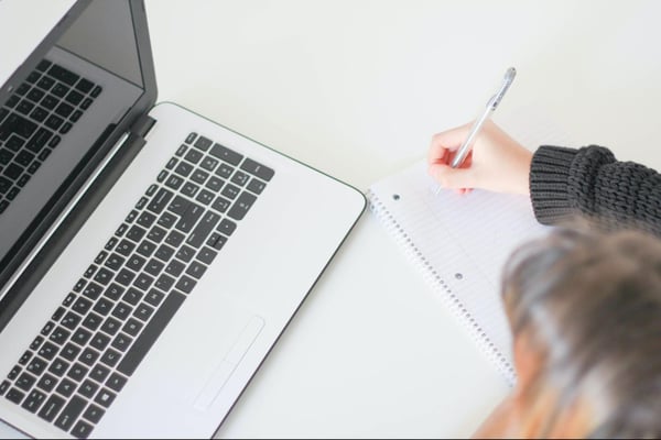 A young person taking notes while using a laptop. 