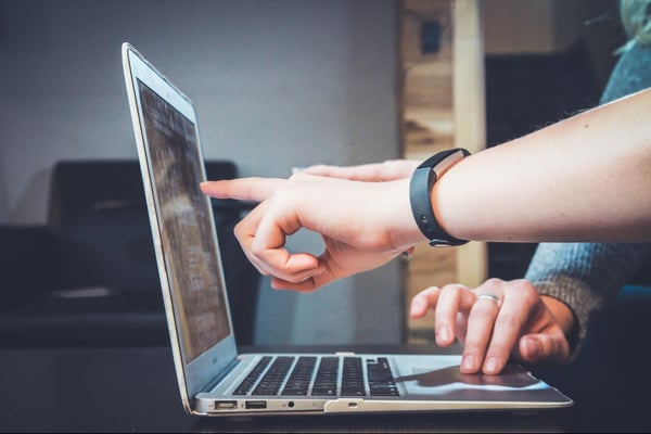 One person’s hand points at a laptop screen while another person’s hand uses the mousepad. 