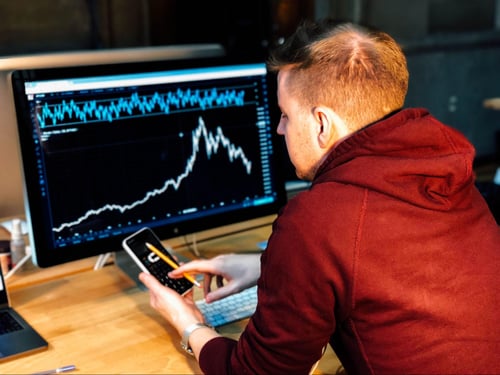 A white man checking his stocks on a desktop computed and also looking at his phone