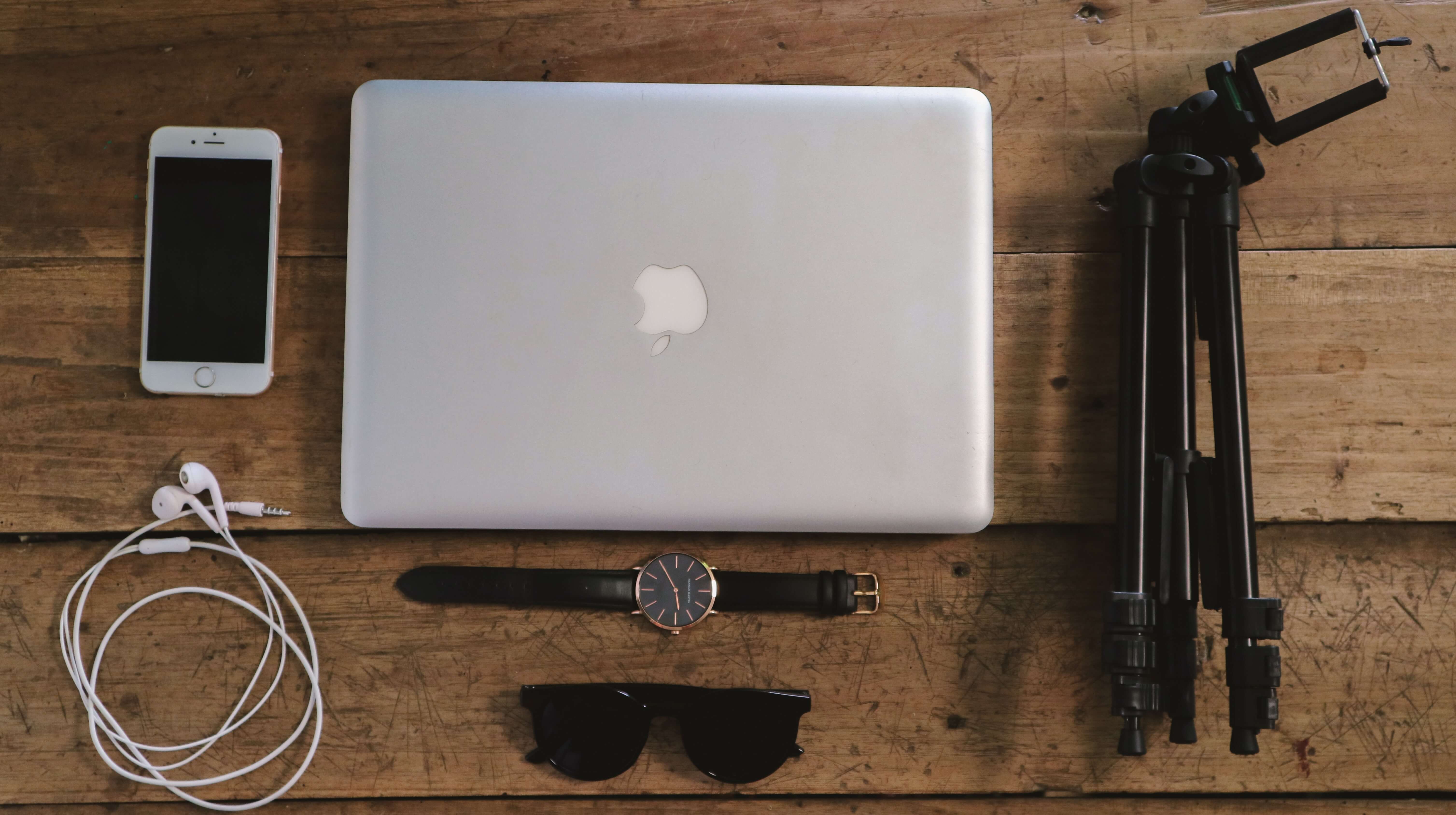 An iPhone, a Macbook, a tripod, a pair of earbud, a watch, and a pair of sunglasses laid out stylishly on a table.