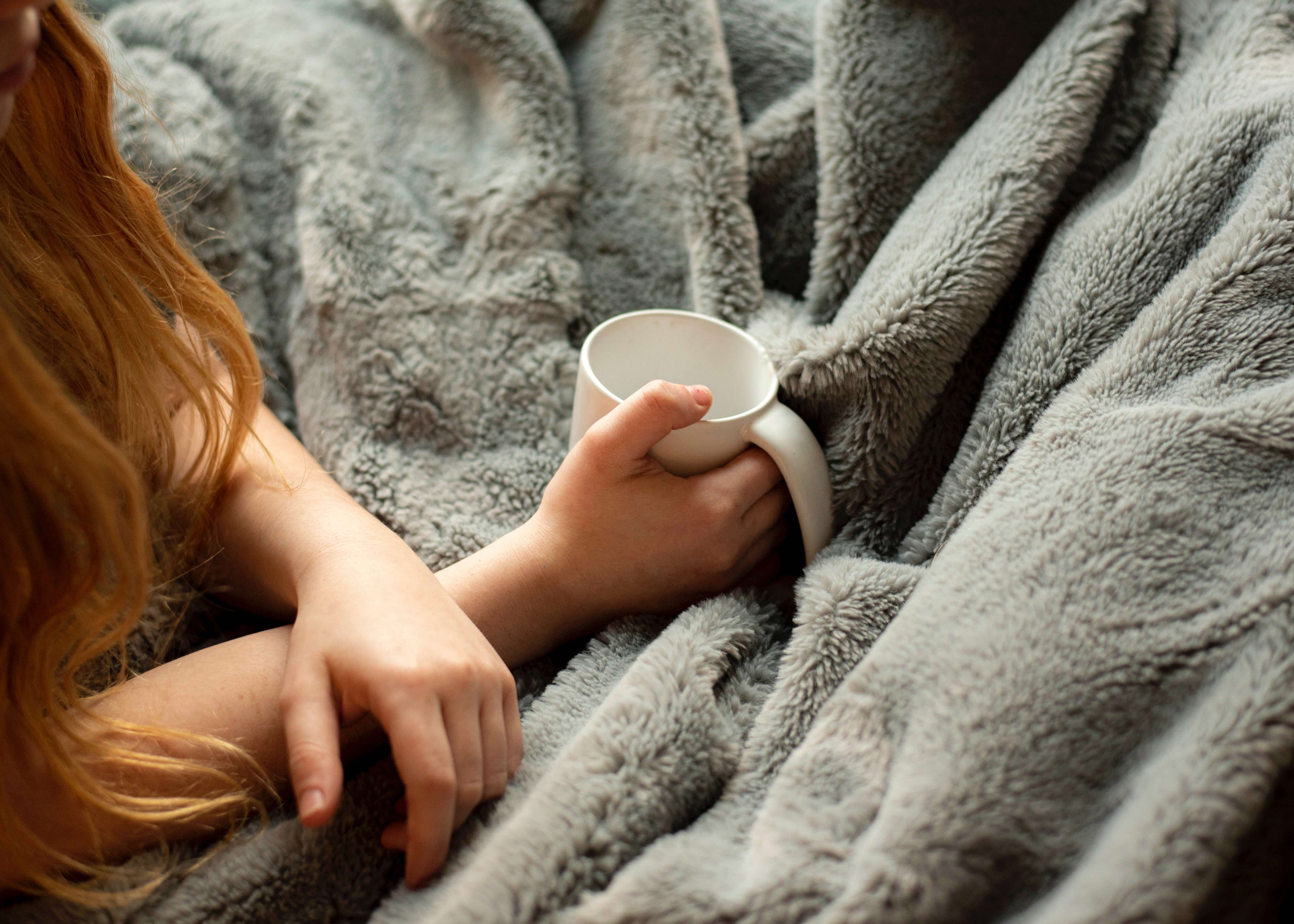 A redhead woman drinks coffee while wrapped in a fuzzy grey blanket. 