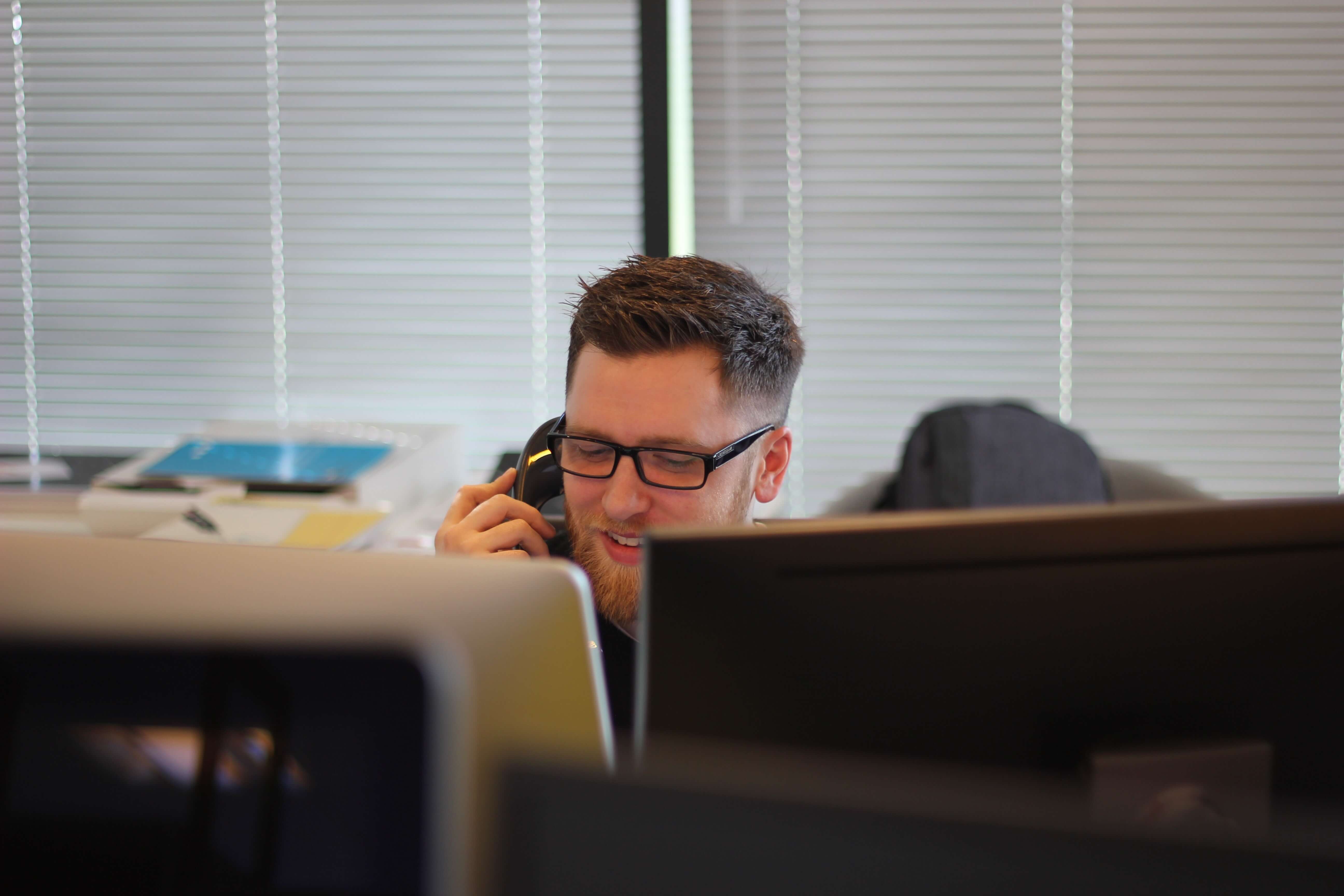 A white man talks on the phone behind two computer monitors