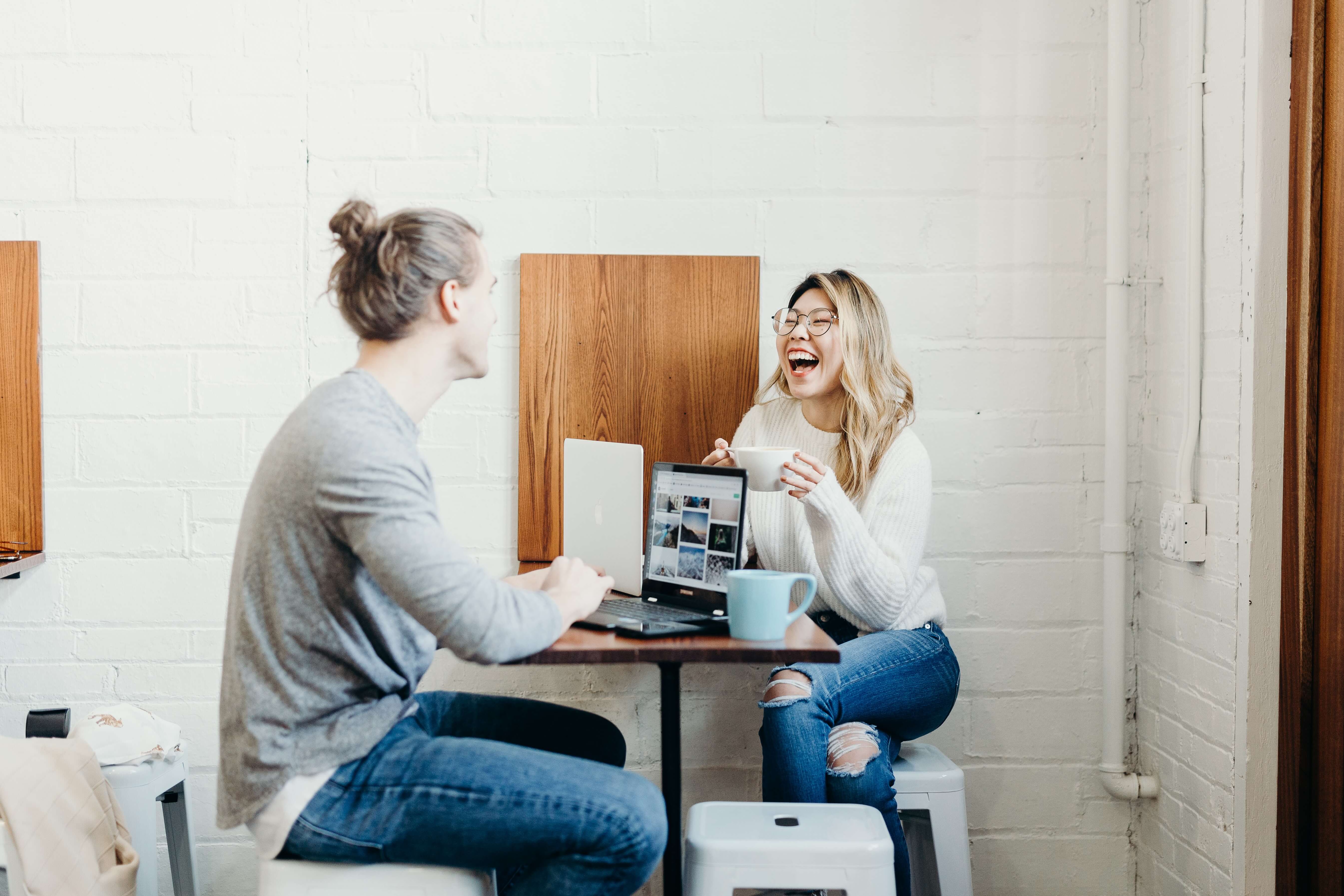 A white man and an east Asian woman each using laptops at a coffee shop. The woman is laughing