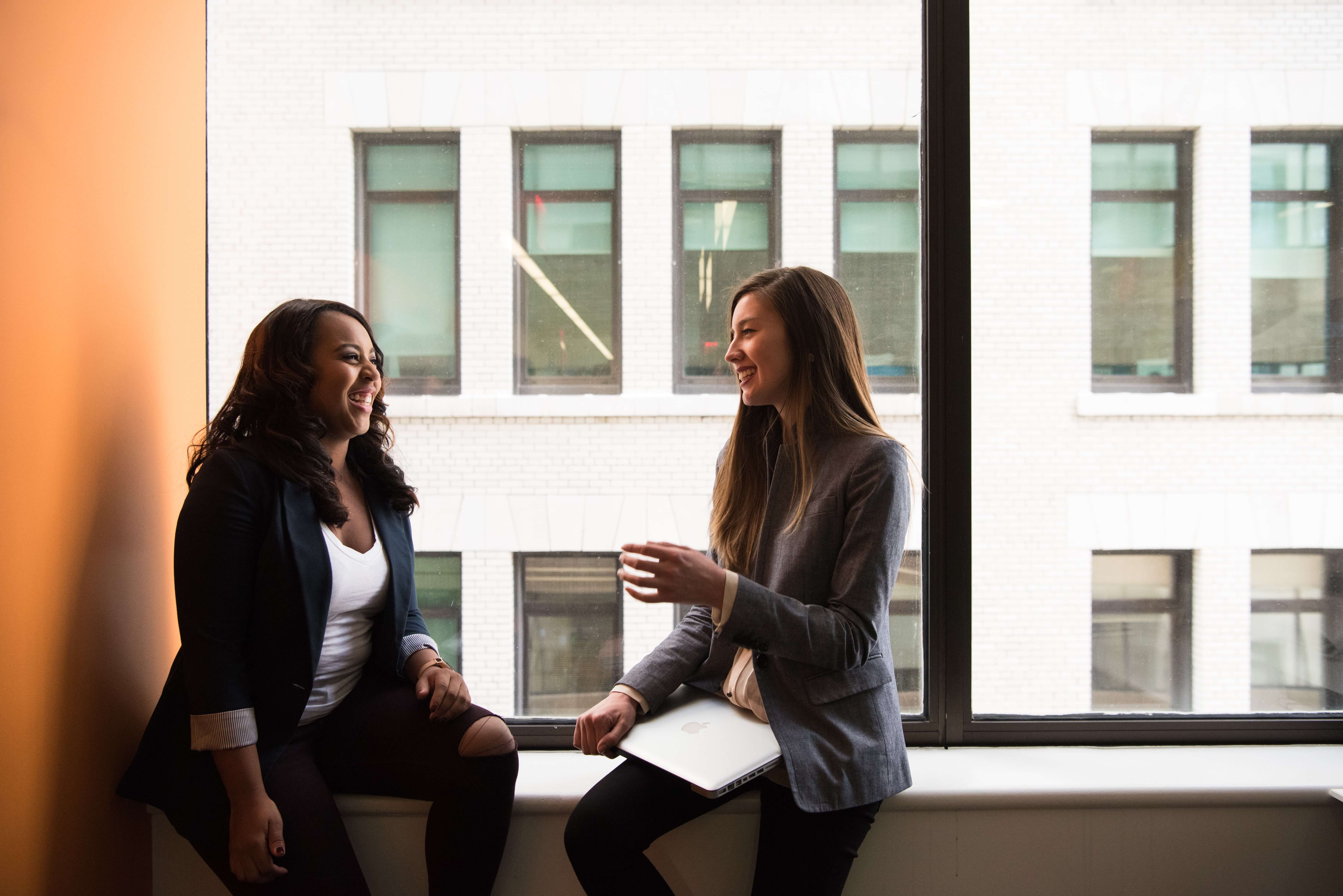 A black woman and a white woman, both in business casual clothing, sitting on a windowsill and having a conversation