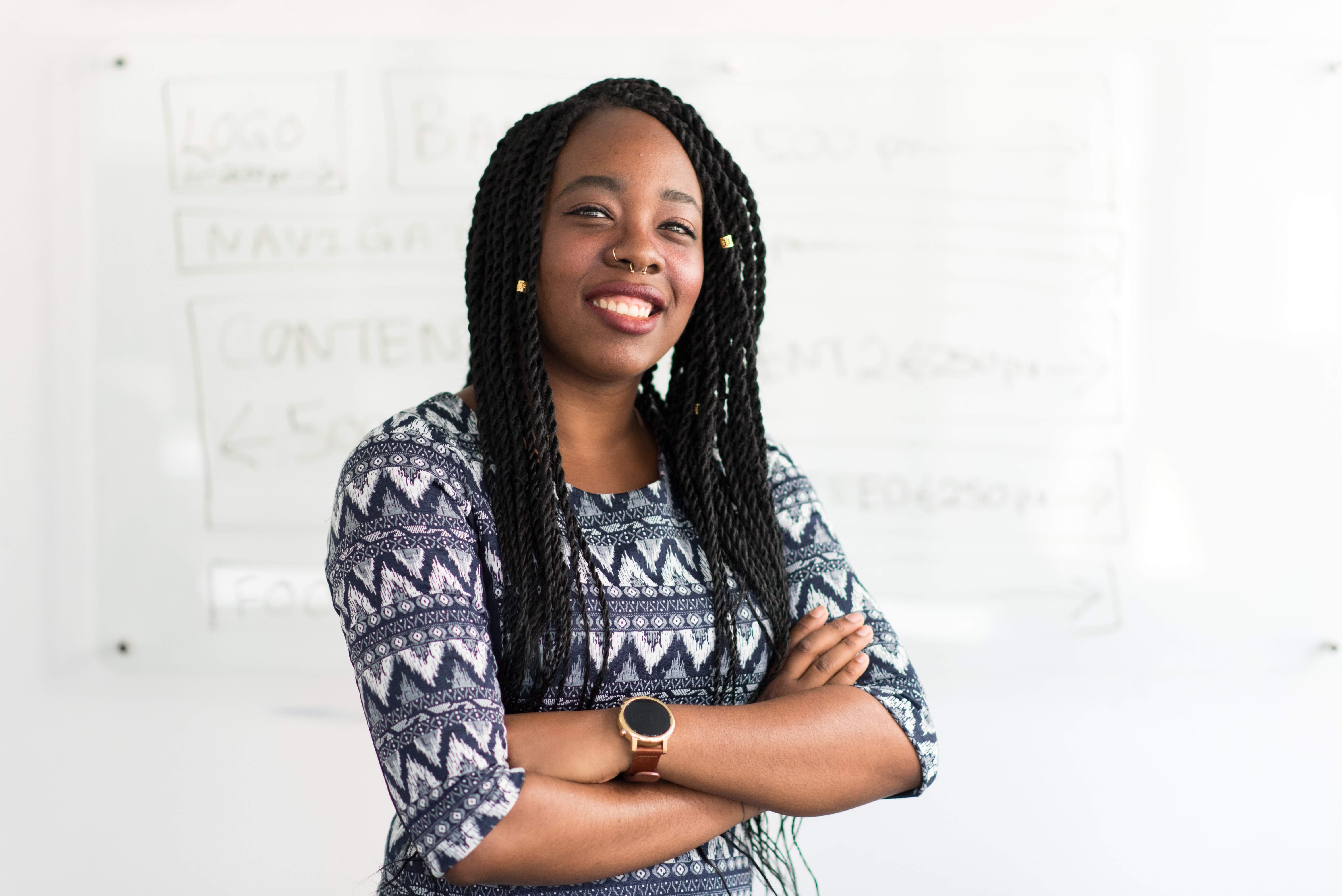 A black woman in business casual clothing smiling and crossing her arms in front of a white board