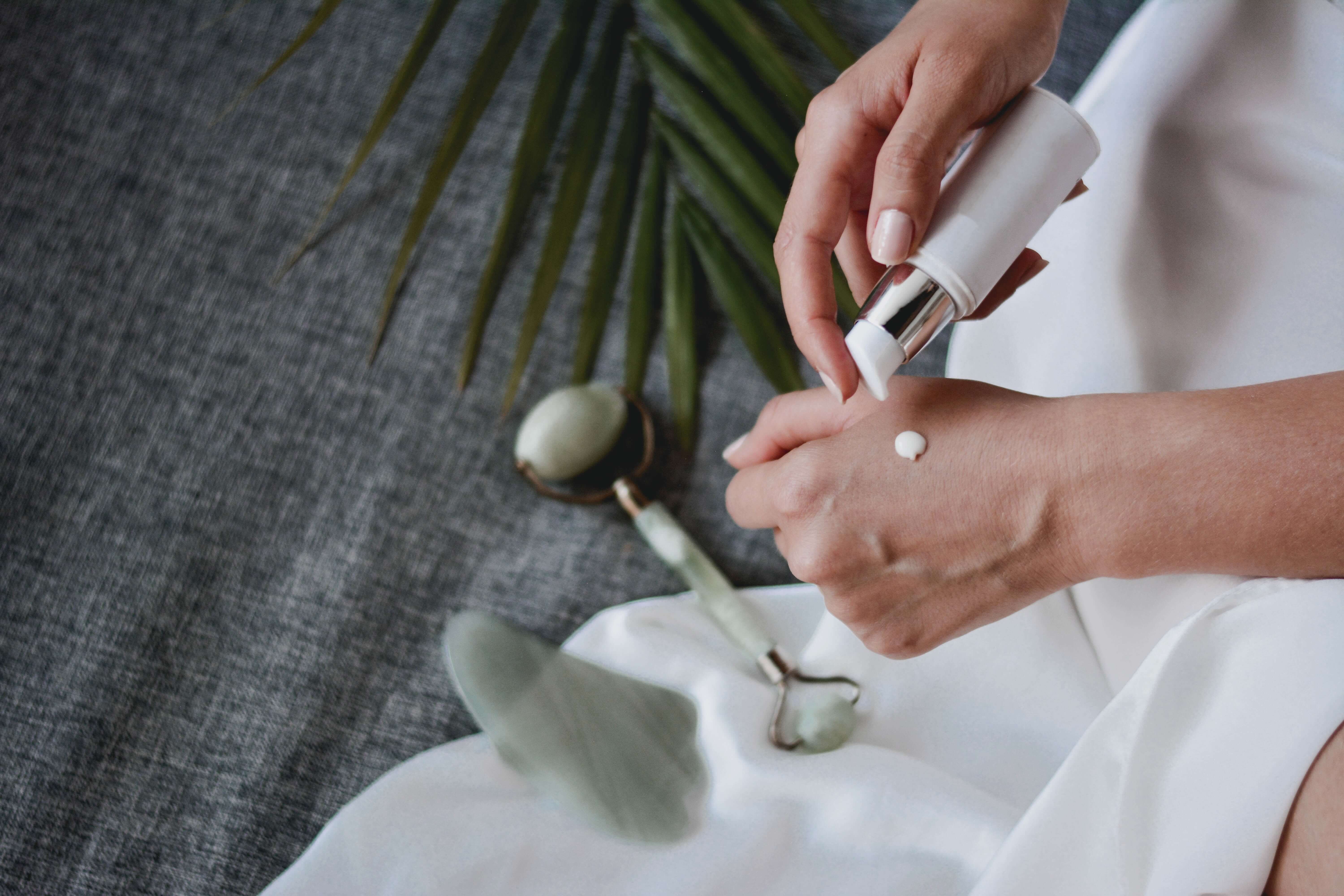 A closeup of a white person putting lotion on their hand. Other skincare products are nearby. 