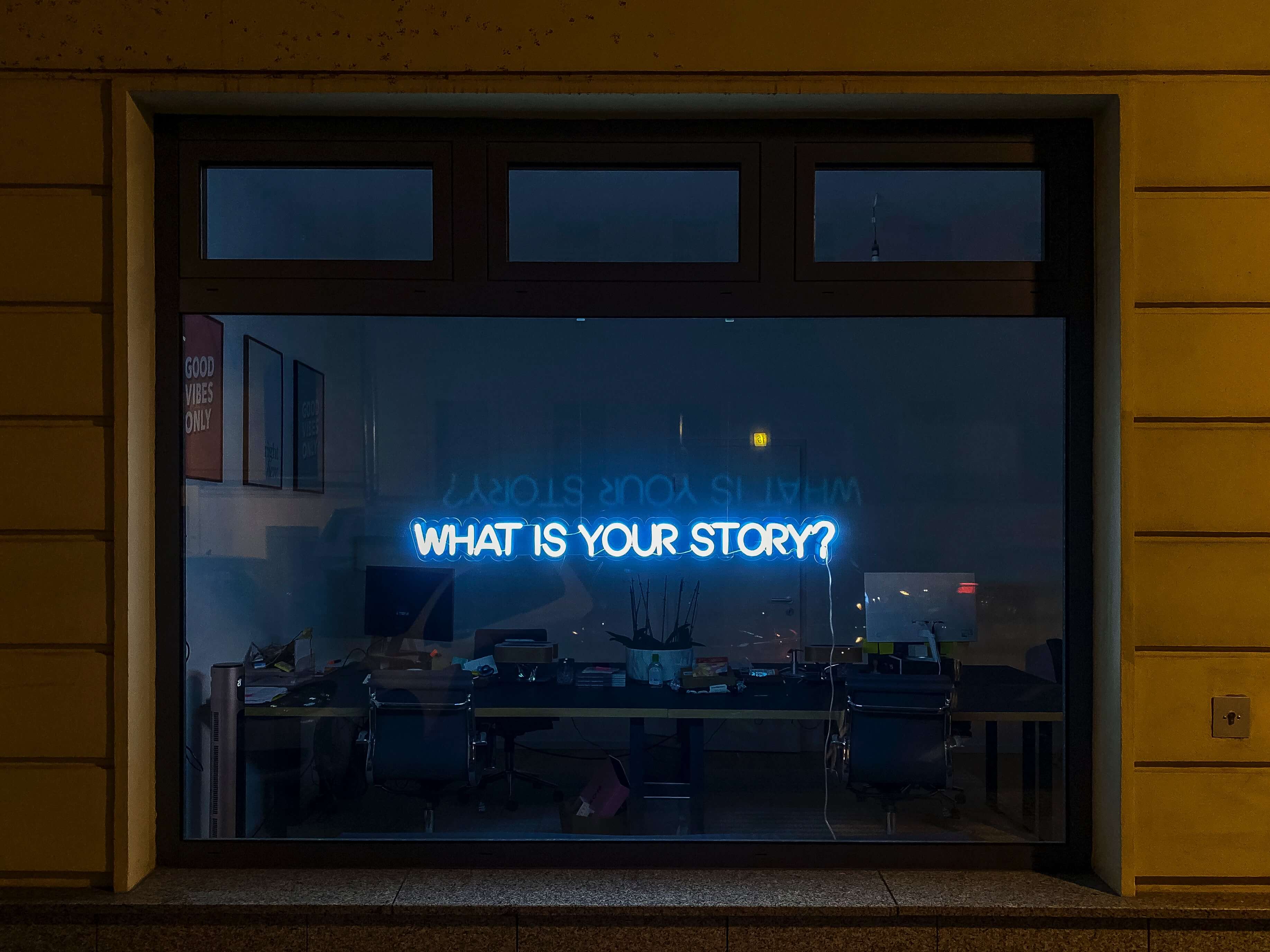 A neon sign that reads "What is your story?" in the window of a darkened office. 