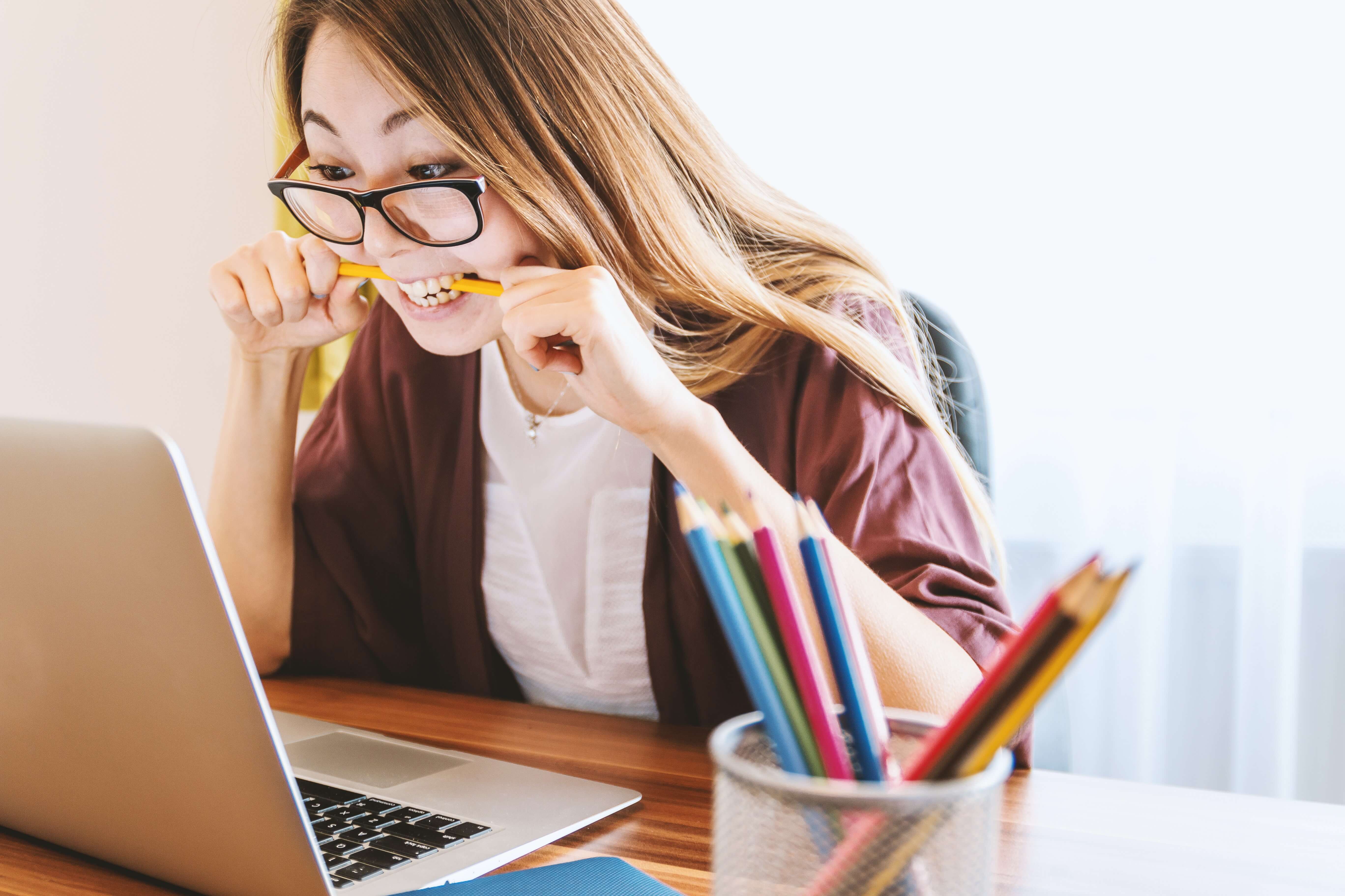 A young East Asian woman wearing glasses bites a pencil while looking at her laptop