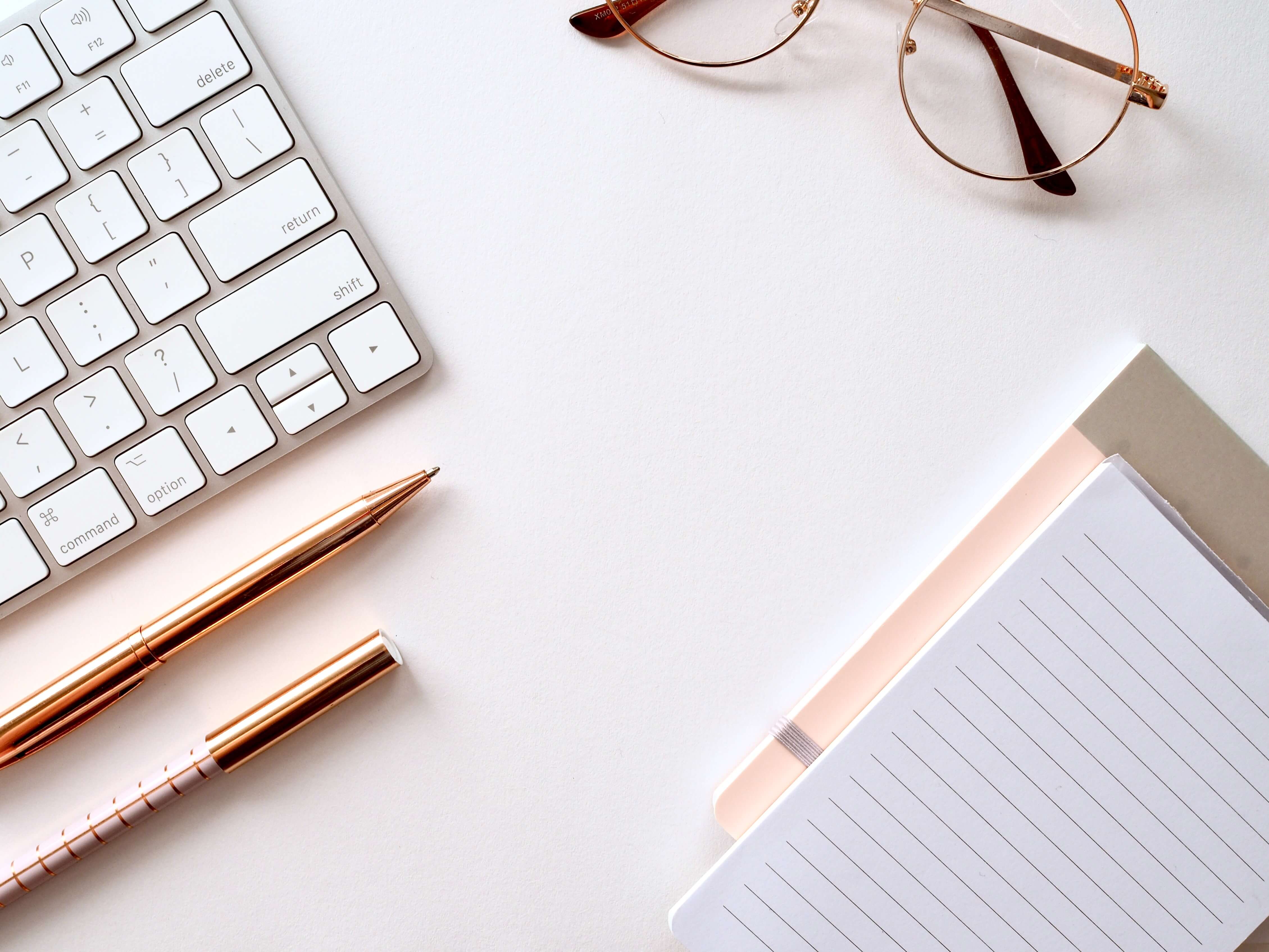 A white desk with a keyboard, two notebooks, two pens, and a pair of glasses. All items are slightly out of frame.