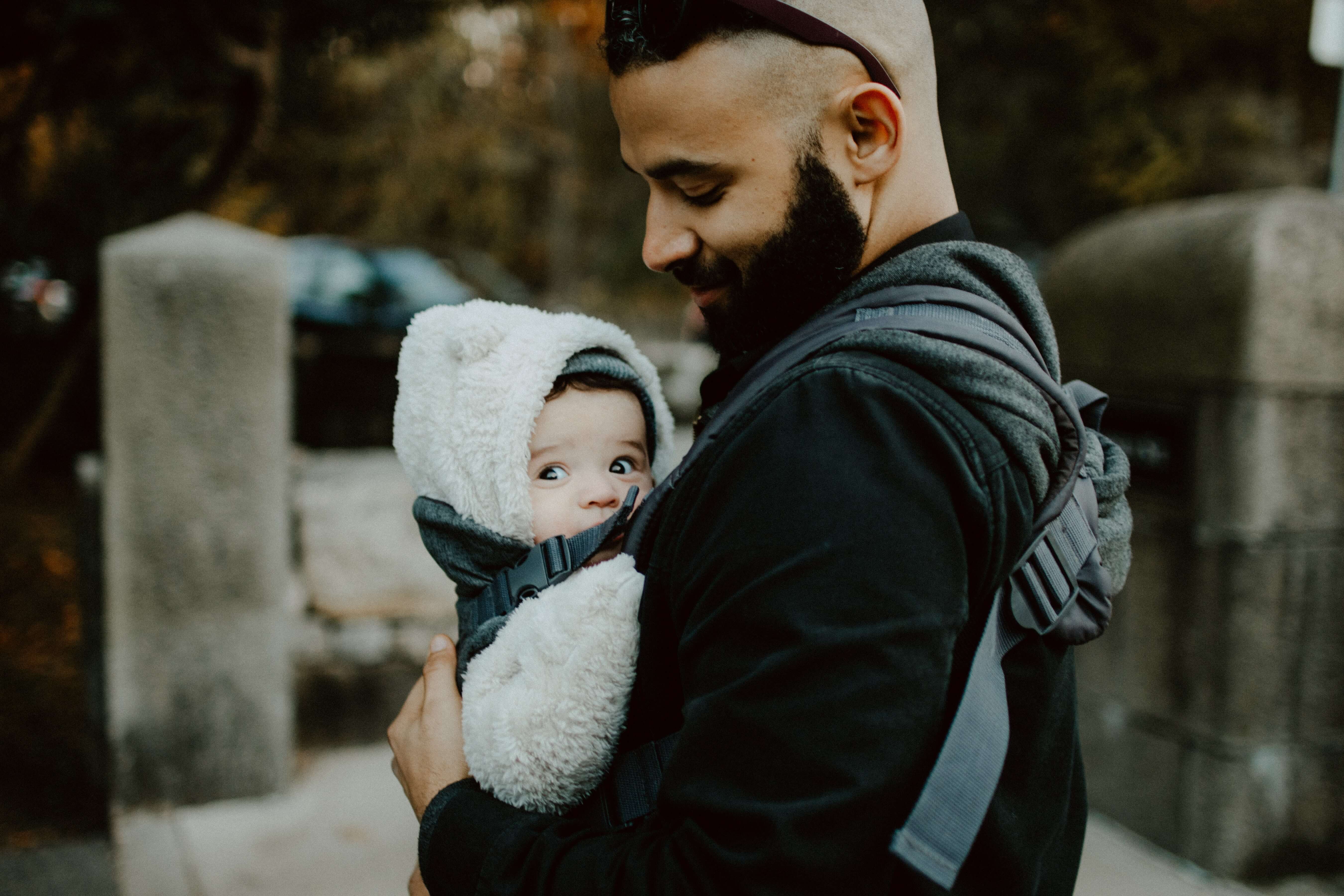 A middle eastern man holds a baby in a fluffy jacket in a front-carrying baby carrier.