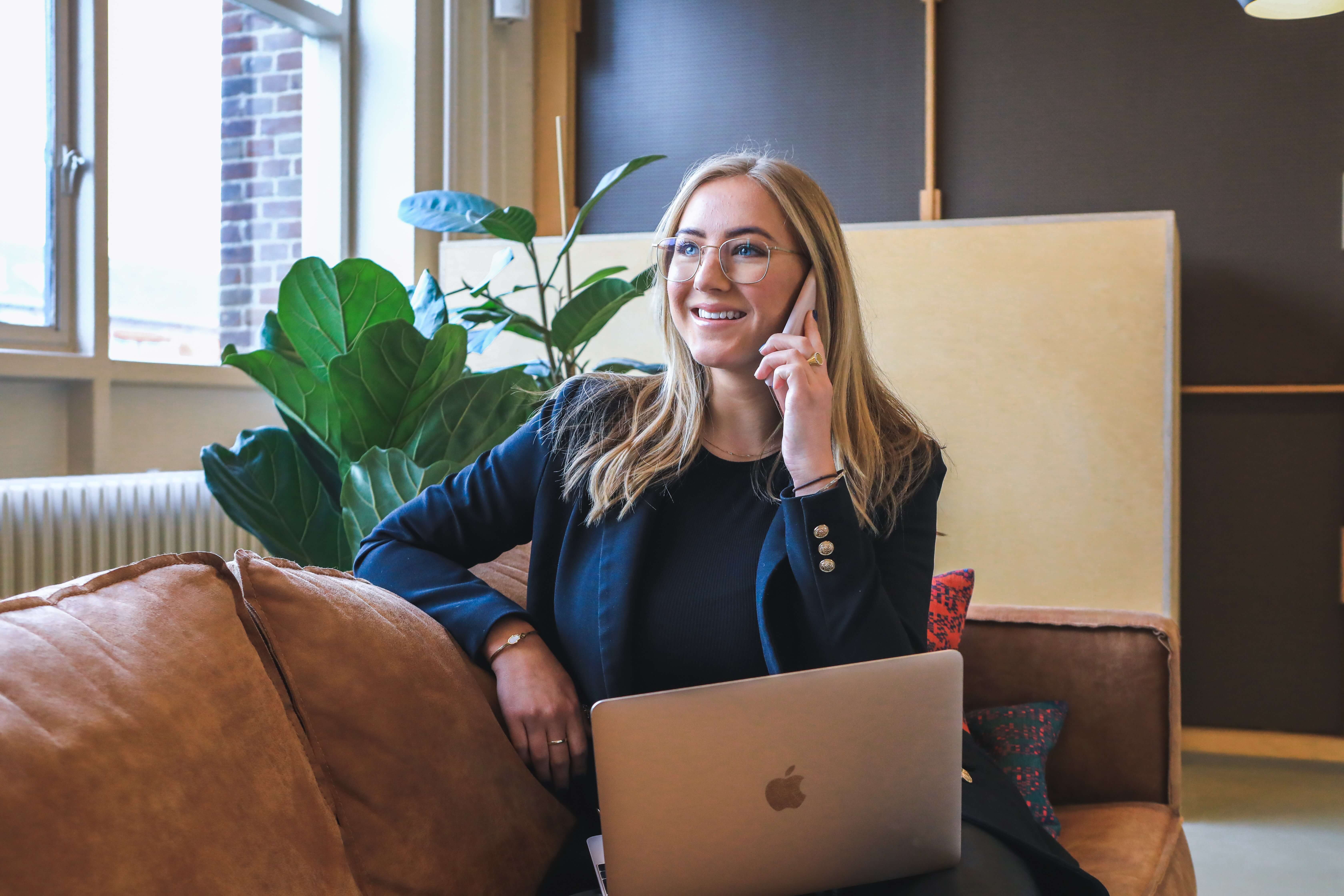 A blonde woman in professional attire talks on the phone while using a laptop