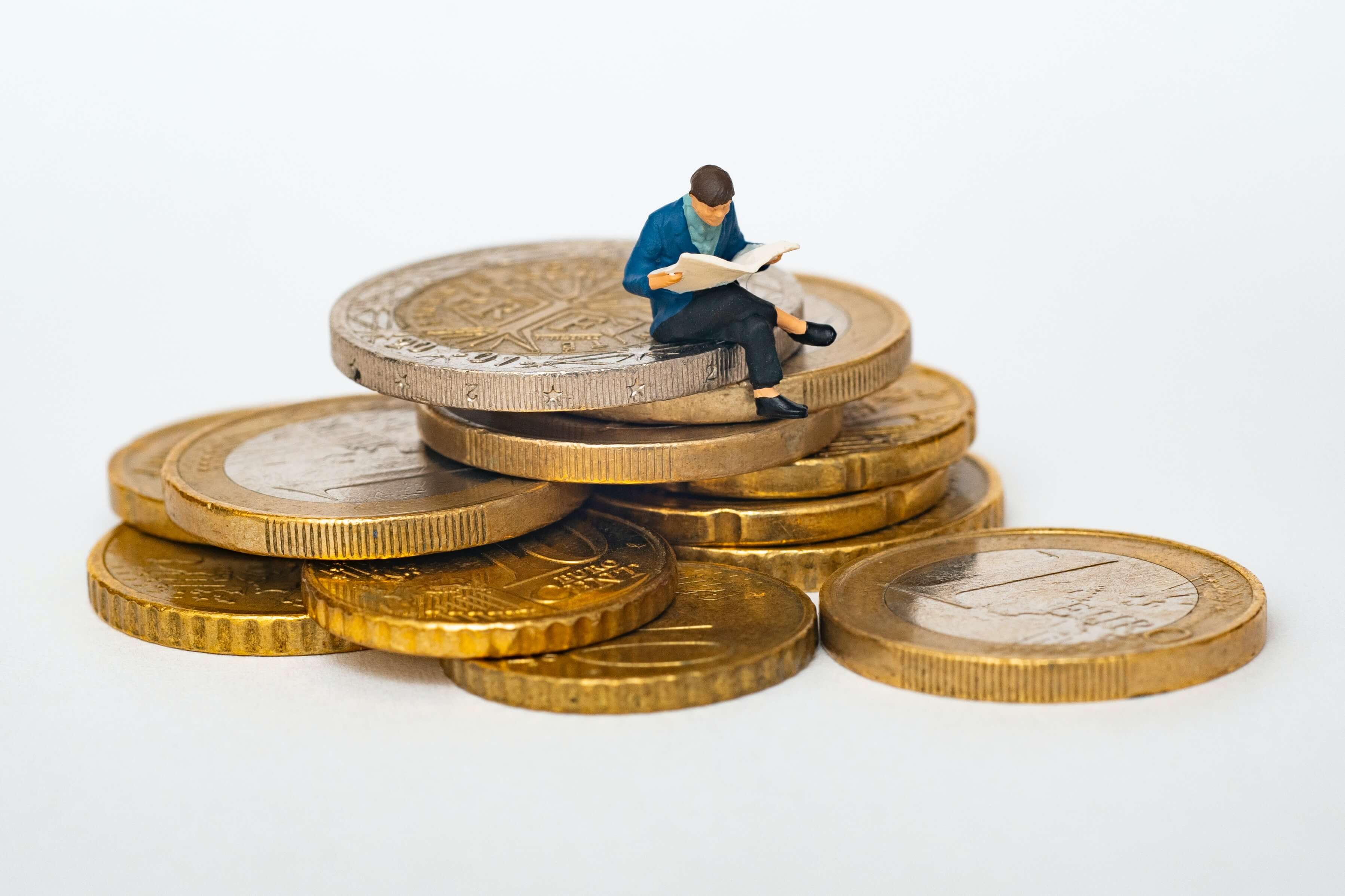 A tiny figurine of a person reading a newspaper sits on a pile of various Euro coins