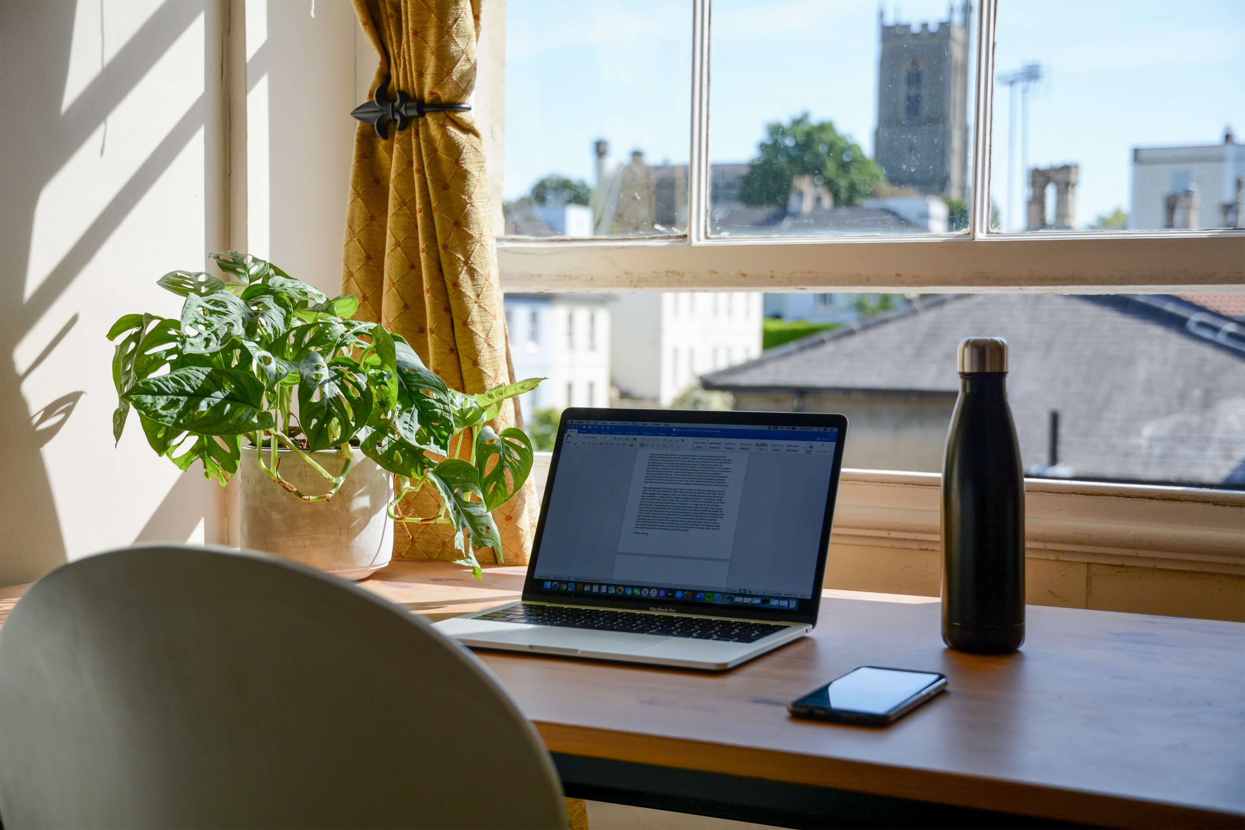 A desk next to an open window with a laptop, water bottle, smartphone, and potted plant set up