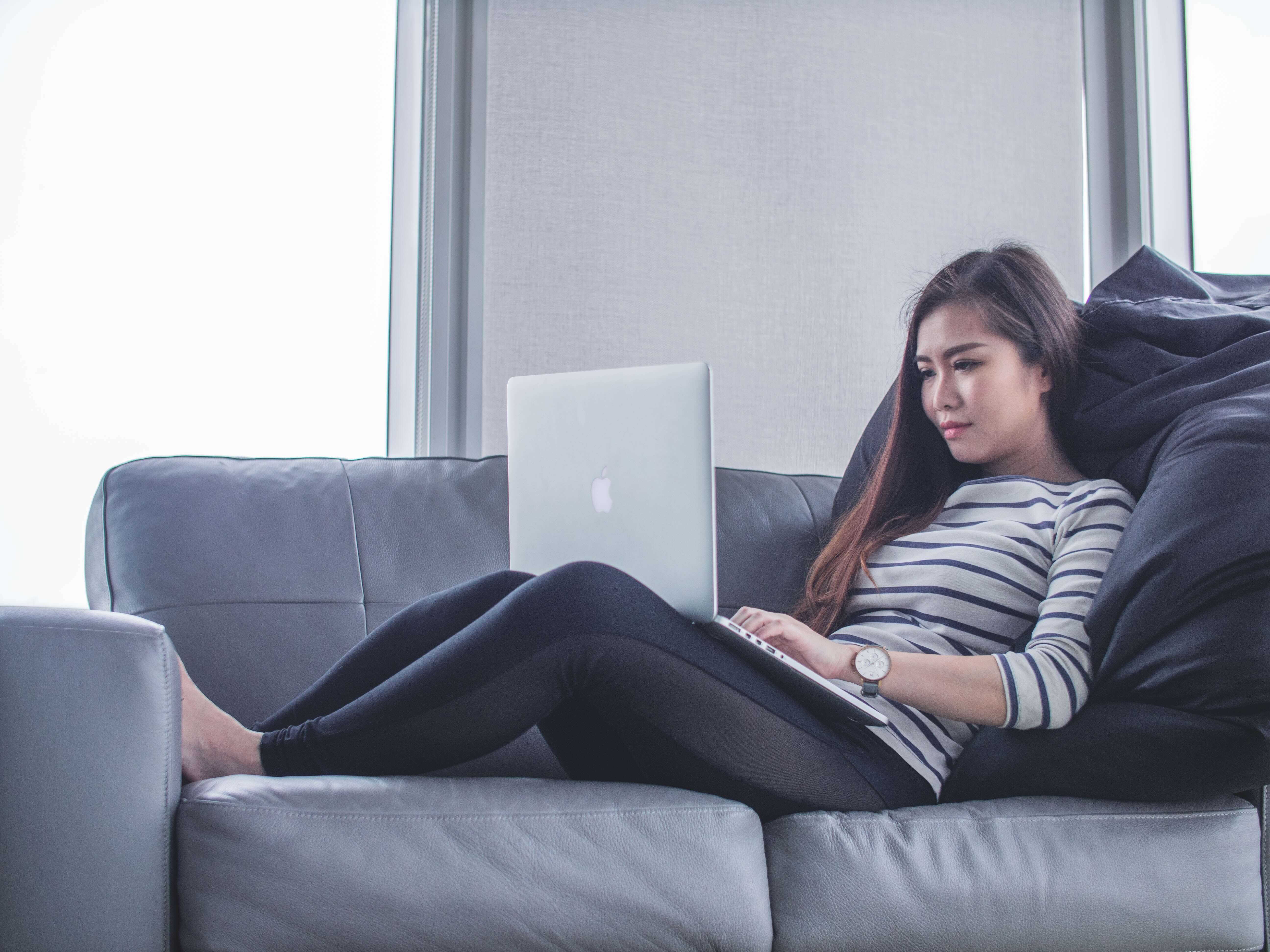An East Asian woman in athleisure clothing  uses a computer while sitting on a grey leather couch