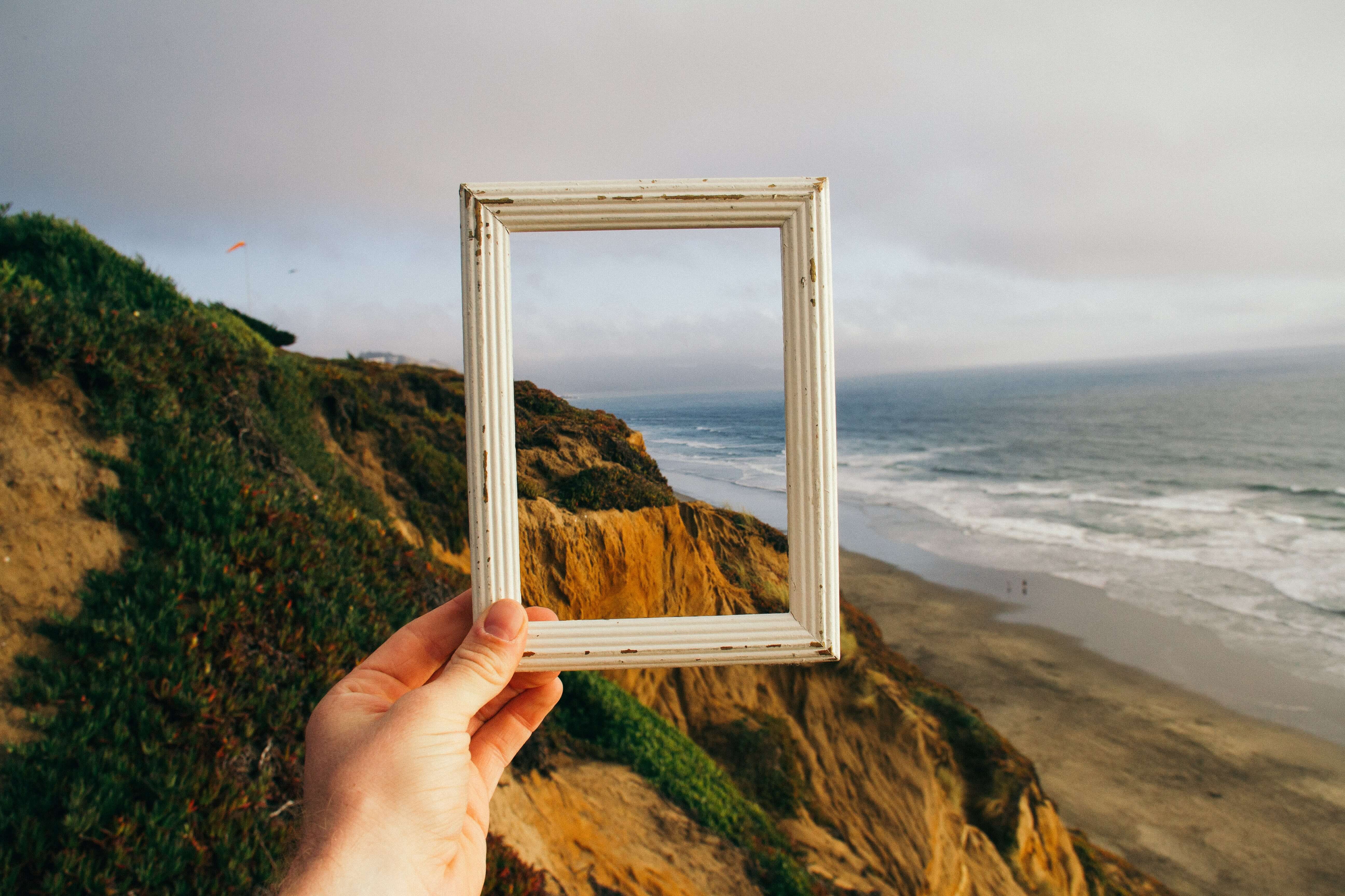  A hand holding up a wooden picture frame around a beachside landscape