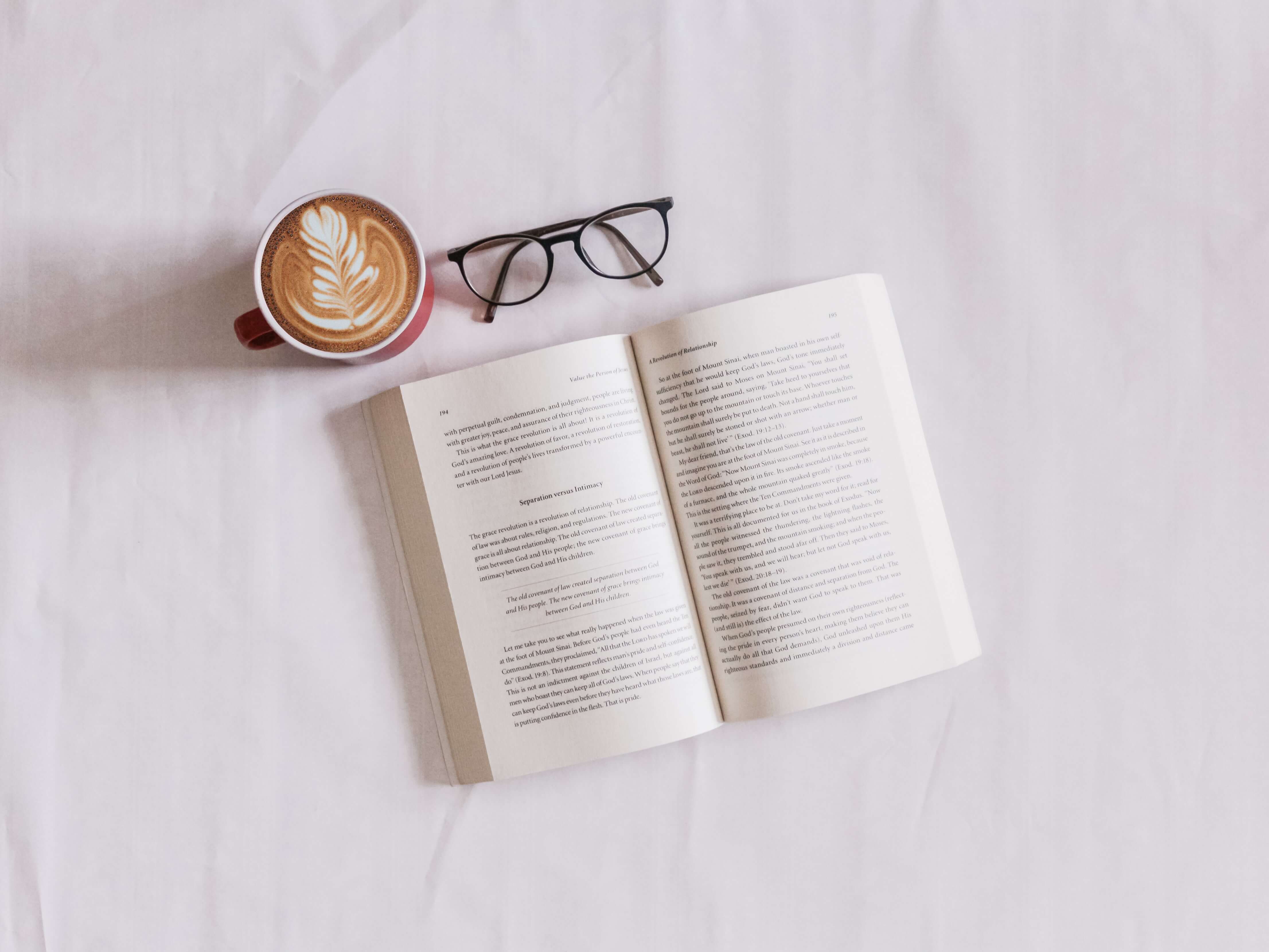 An open book, a pair of glasses, and a latte with a fern drawn on the top with foam on a white background