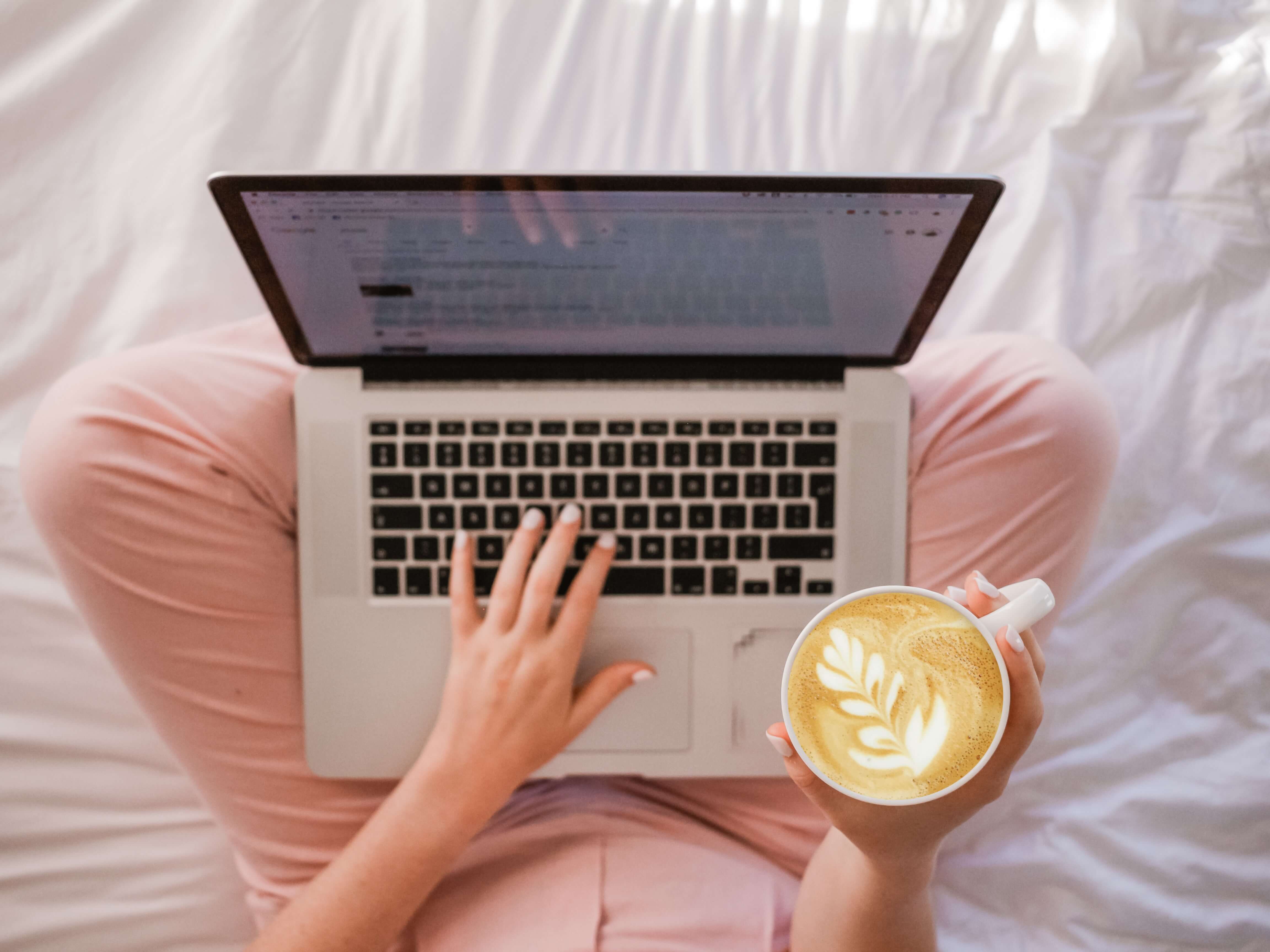 A shot from above of a person sitting on a bed using a laptop while holding a cappuccino with fancy foam art on it