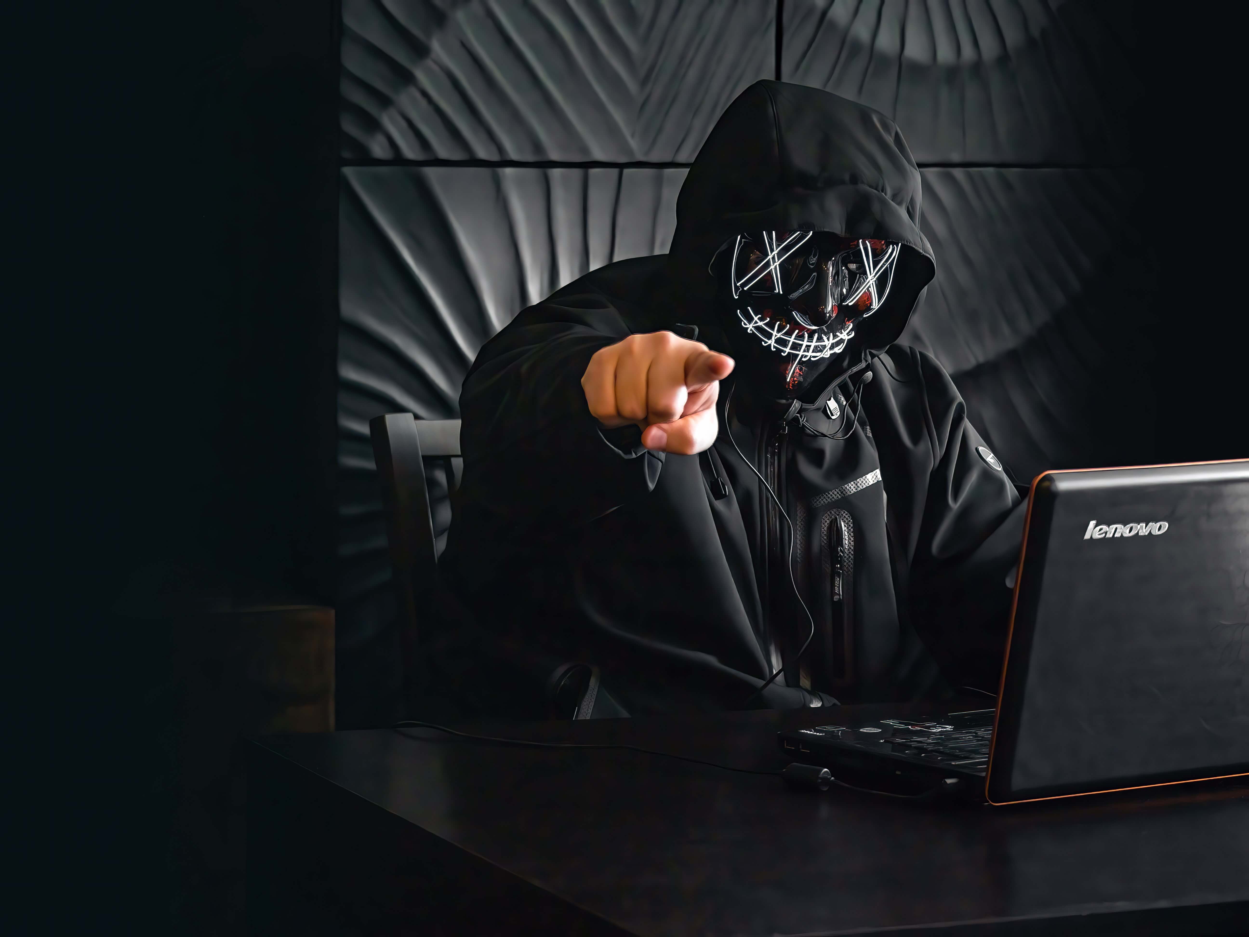 A person in a hacker mask using a laptop and pointing towards the camera