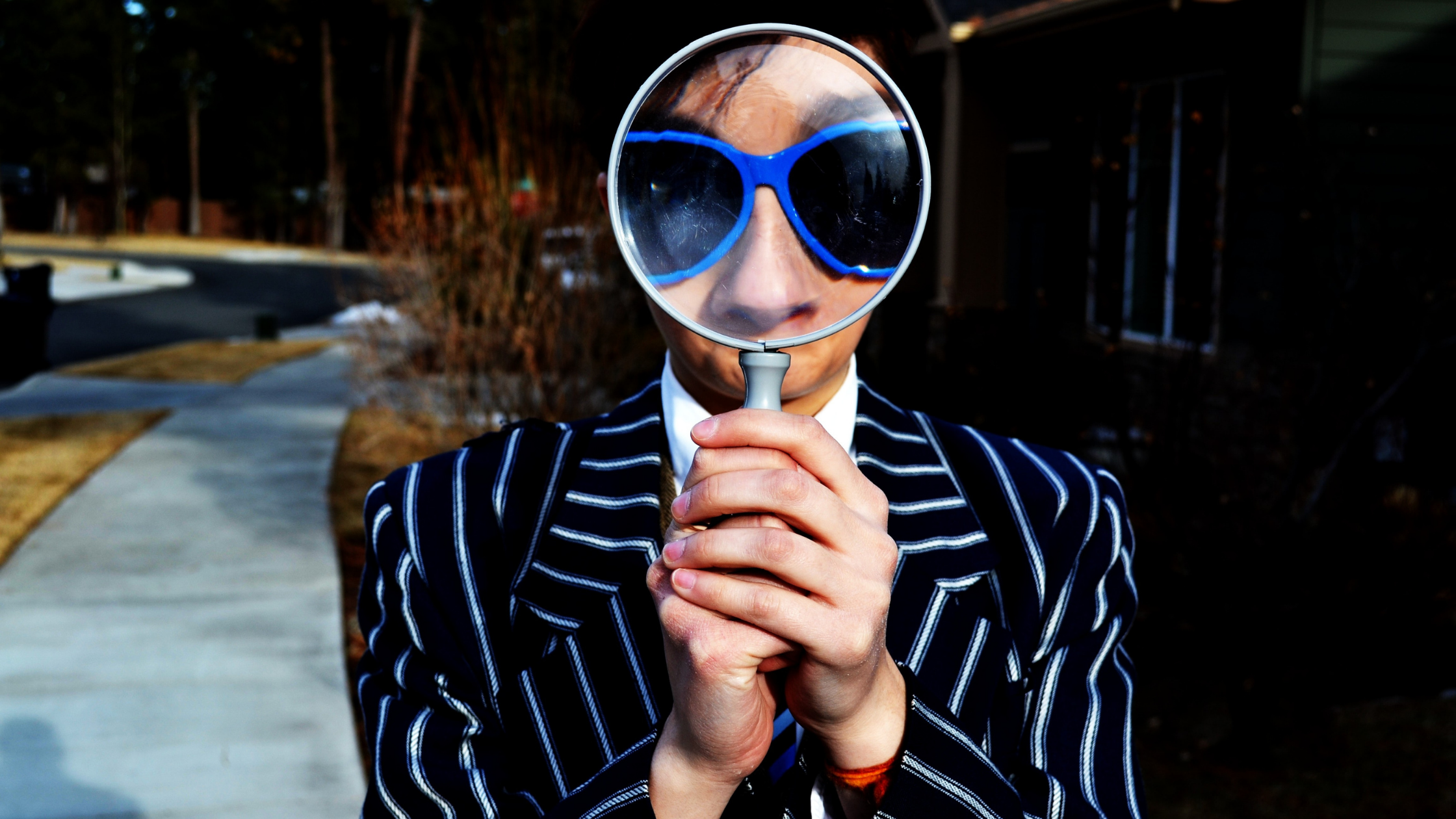 A person in a pinstriped suit and sunglasses holds a magnifying glass up to their face
