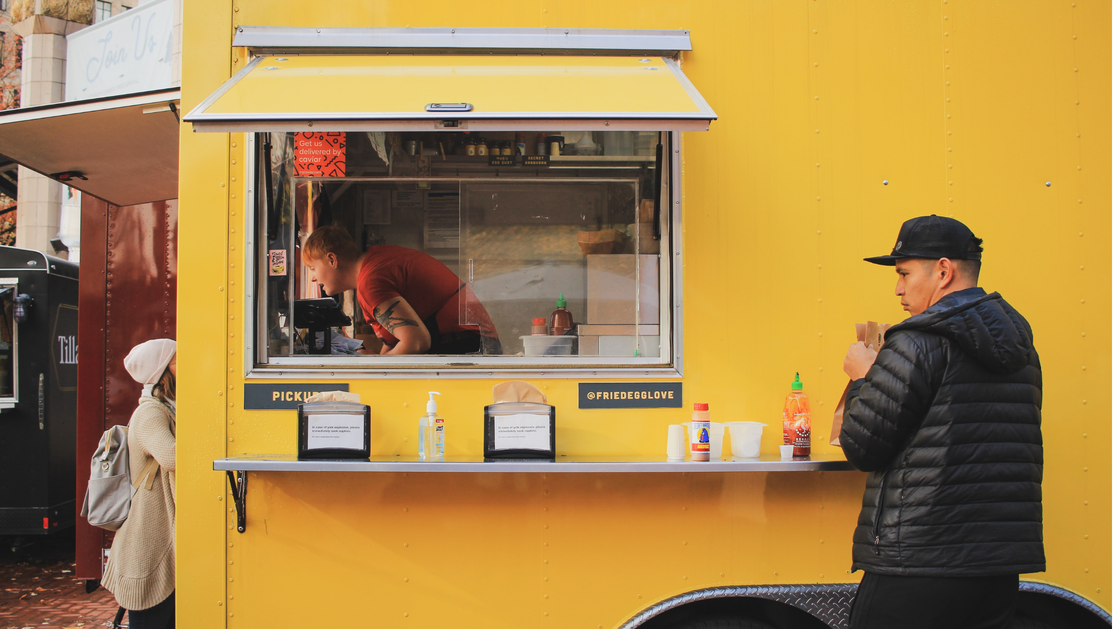 A customer in a black jacket and hat outside a yellow food truck